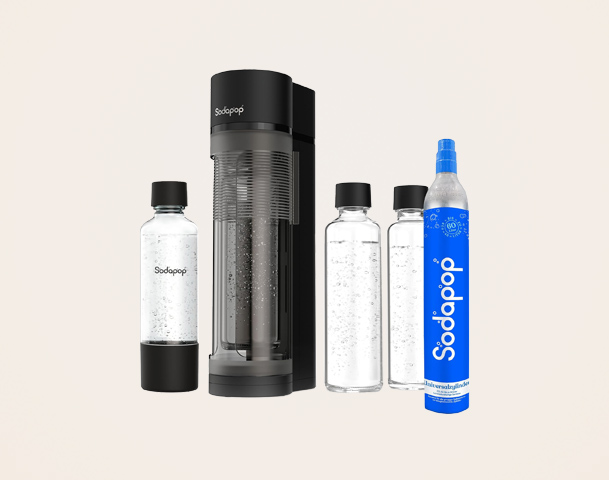 Das Bild zeigt ein Sodapop-Gerät zur Herstellung von Sprudelwasser. Es sind vier Wasserflaschen zu sehen: eine mit deutlich sichtbarem Sodapop-Logo und drei weitere neben dem Sprudler. Der Hintergrund ist hell und neutral gehalten.