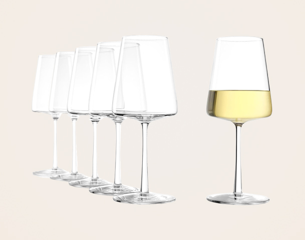 Sechs leere Weingläser stehen in einer Reihe, während ein einzelnes Glas mit weißem Wein gefüllt ist, das sich leicht abhebt. Der Hintergrund ist hell und neutral, um den Fokus auf die Gläser zu lenken.