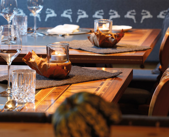 Der Tisch ist liebevoll gedeckt mit Weingläsern und einem Kerzenhalter aus Glas, der in einem Blatt sitzt. Eine graue Tischdecke und elegante Bestecke ergänzen das Ambiente. Der Hintergrund zeigt eine stilvolle Wandgestaltung.