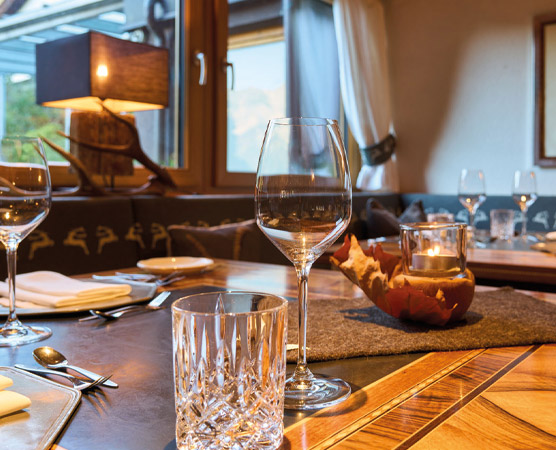 Ein elegant gedeckter Tisch in einem Restaurant, mit mehreren Gläsern, Besteck und einem kleinen Kerzenhalter. Ein weicher Teppich und Fenster geben einen Blick auf die Außenlandschaft. Das warme Licht schafft eine einladende Atmosphäre.