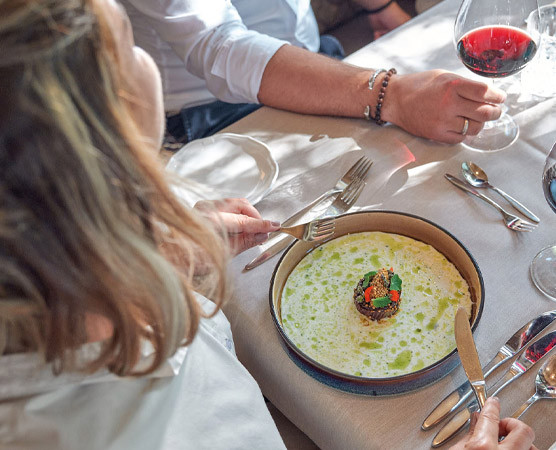 Eine Person sitzt an einem Tisch und betrachtet einen kunstvoll angerichteten Teller mit einer Speise. Die garnierte Speise ist von einer hellen, schaumigen Sauce umgeben. Neben dem Teller stehen Besteck und ein Glas Rotwein.