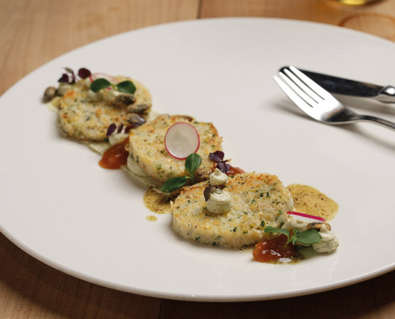 Ein weißer Teller mit drei goldbraun gebratenen, runden Patties, garniert mit kleinen grünen Blättchen, Radieschen und einer dunklen Sauce. Neben den Patties befinden sich dekorative Tropfen von einer fruchtigen Sauce. Ein Besteck aus Metall liegt am Rand des Tellers.