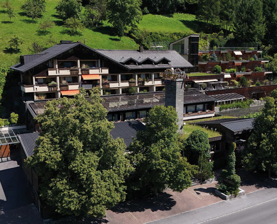 Ein modernes Hotelgebäude mit mehreren Etagen, umgeben von Bäumen und grünen Wiesen. Das Dach hat eine charakteristische Form, und Balkone sind mit Pflanzen dekoriert. Im Vordergrund befindet sich eine Straße, die zum Hotel führt.