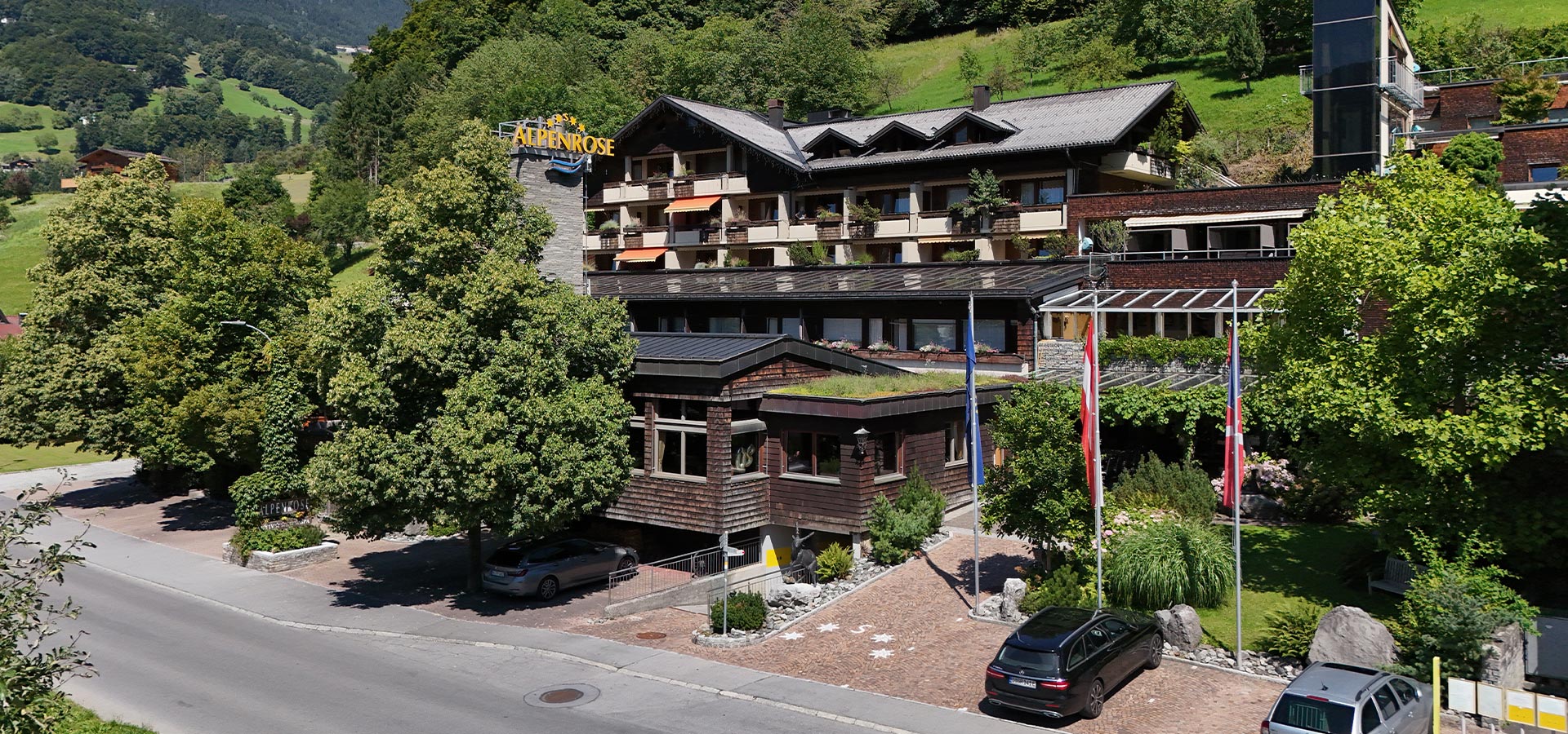 Ein modernes Hotelgebäude mit Holzverkleidung und mehreren Stockwerken, umgeben von grünen Bäumen und Wiesen. Vor dem Hotel stehen Autos geparkt. Im Hintergrund sind sanfte Hügel zu sehen. Ein Schild mit der Aufschrift „Alpenrose“ ist sichtbar.