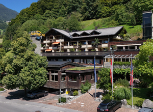 Ein modernes Hotelgebäude mit Holzverkleidung, umgeben von grünen Bäumen und Wiese. Im Vordergrund sind Flaggen aufgestellt. Das Hotel hat mehrere Balkone und ist von einer malerischen Landschaft umgeben.
