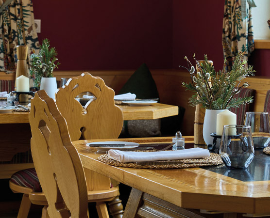 Ein gemütlicher Gastraum mit Holztafeln und detaillierten Stühlen. Auf den Tischen stehen dekorative Pflanzen in Vasen, Kerzen und Gläser. Die Wände sind in einem warmen Rotton gehalten, mit Fenstern, die mit gemusterten Vorhängen geschmückt sind.
