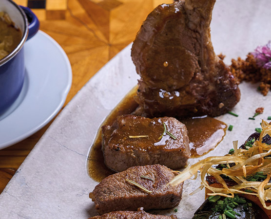 Ein Teller mit zartem Fleisch, darunter Stücke von Rindersteak und ein Lammkotelett, serviert mit einer reichhaltigen Sauce. Dazu sind knusprige Zwiebelstreifen und Beilagen auf dem Tisch angerichtet. Der Hintergrund zeigt eine ansprechend gestaltete Tischplatte.