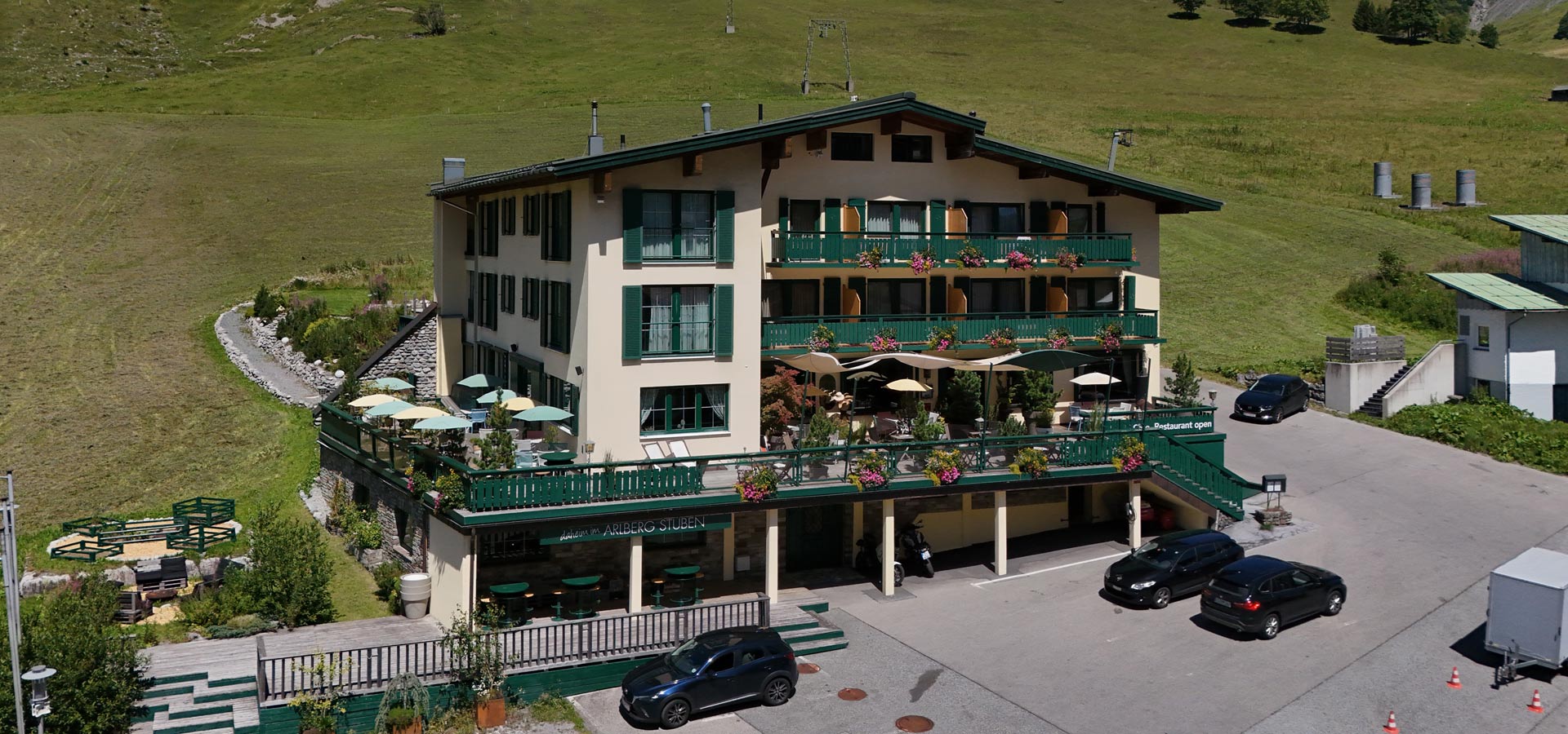 Ein mehrstöckiges Gebäude mit grünem Balkon und blühenden Pflanzen. Vor dem Gebäude stehen Tische mit Sonnenschirmen. Parkplätze sind im Vordergrund sichtbar. Die Umgebung besteht aus einer grünen Wiese mit sanften Hügeln im Hintergrund.