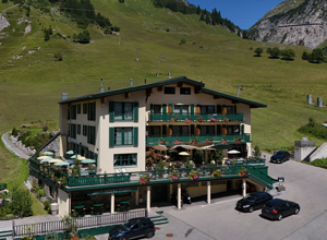 Das Bild zeigt ein mehrstöckiges Hotelgebäude in einer gebirgigen Landschaft. Es hat grüne Balkone und eine Terrasse mit Sonnenschirmen. Vor dem Hotel sind mehrere Autos geparkt, und die Umgebung ist von grünen Wiesen und Bergen umgeben.