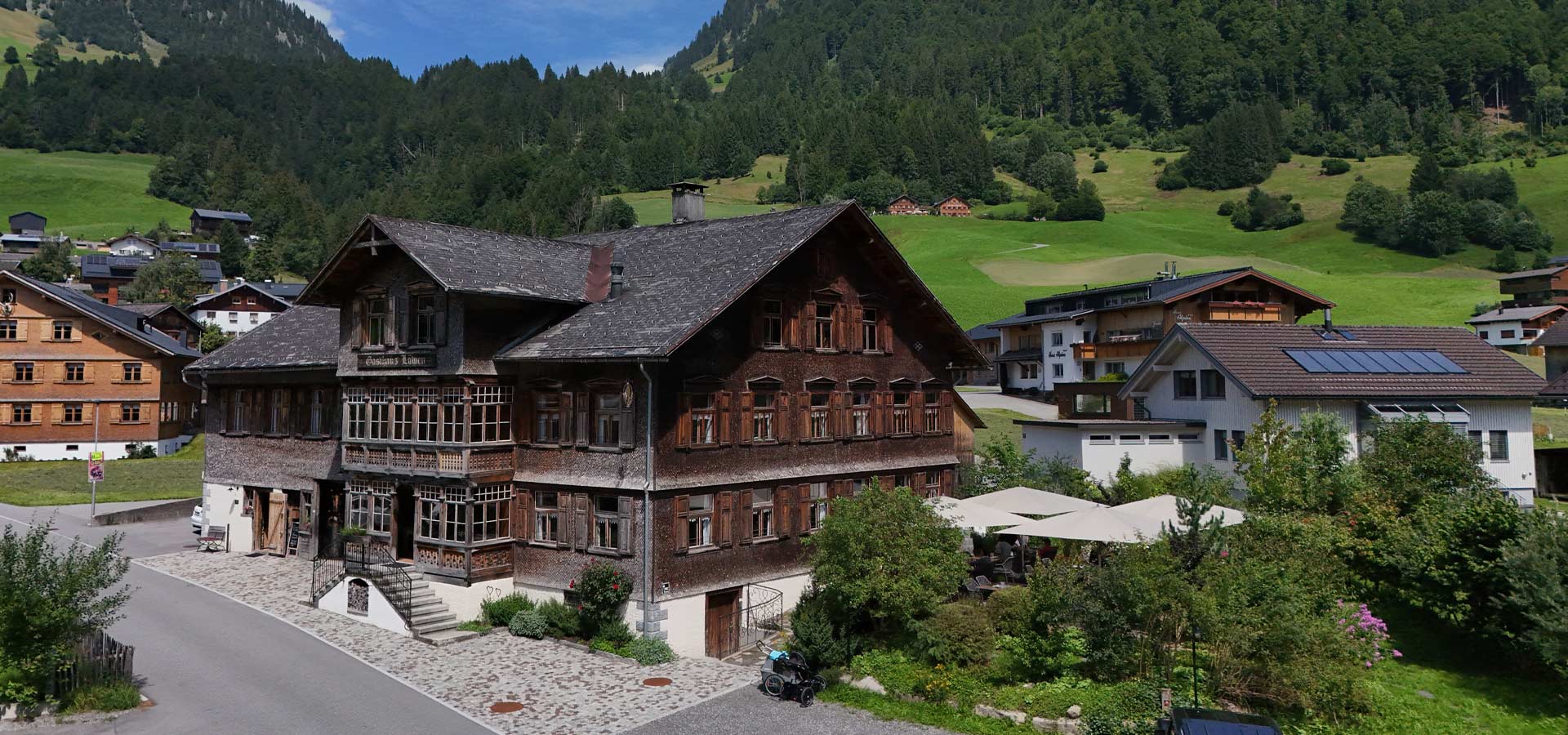 Ein traditionelles Holzhaus mit vielen Fenstern und einem großen, begrünten Garten im Vordergrund. Im Hintergrund sind sanfte Hügel und weitere Gebäude zu sehen. Der Himmel ist klar und sonnig.