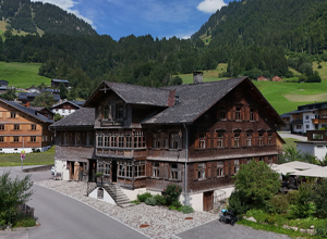 Ein großes, traditionelles Holzhaus steht an einer Straße, umgeben von grünen Wiesen und Bergen. Die Fassade ist reich mit Fenstern und Balkonen verziert. Im Vordergrund sind Pflanzen und ein Weg zu sehen. Die Szenerie vermittelt ein Gefühl von ländlicher Idylle.