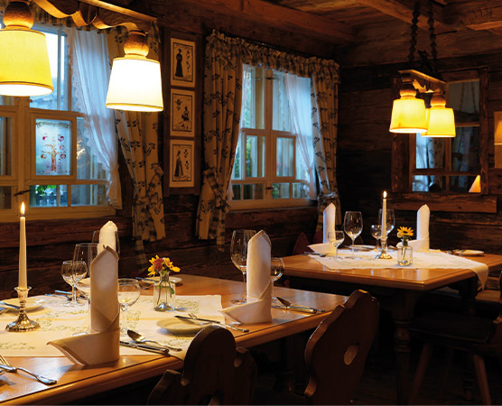 Ein gemütliches Restaurant mit Holzwänden, sanfter Beleuchtung und Tischen, die mit weißen Tischdecken eingedeckt sind. Auf den Tischen stehen Gläser, Besteck und kleine Blumenarrangements. Kerzen sorgen für eine behagliche Atmosphäre. Vor den Fenstern hängen Vorhänge, und an Wänden sind Bilderrahmen mit dekorativen Motiven.