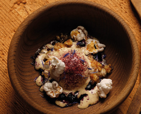 Eine Holzschüssel enthält ein kreatives Gericht mit einer cremigen, weißen Sauce, Toppings aus Popcorn, und einer dunklen, fruchtigen Basis. Die Speise ist mit essbaren Blüten und buntem Pulver garniert, was ihr ein ansprechendes Aussehen verleiht.