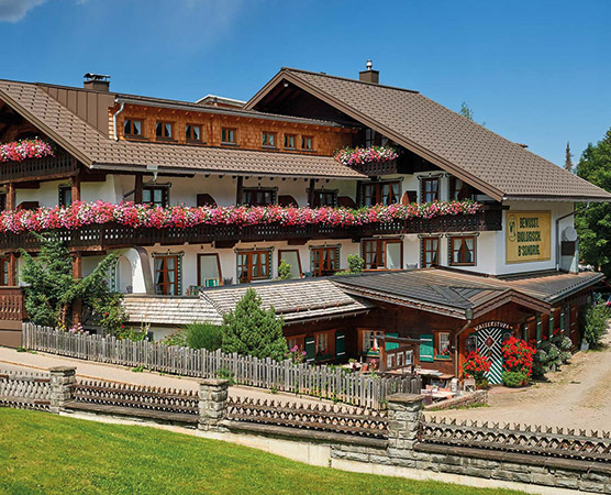 Ein traditionelles alpenländisches Gasthaus mit einem schönen Holzdesign. Der Balkon ist mit bunten Blumen dekoriert, und der Eingang ist von einem grünen Rasen umgeben. Die Fassade strahlt eine einladende Atmosphäre aus, typisch für Bergregionen.
