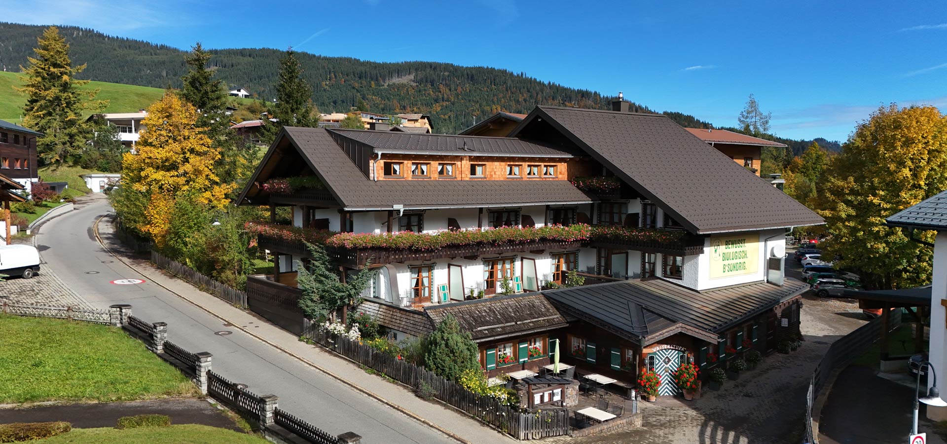 Ein malerisches Alpenhaus mit holzverkleideten Fenstern und bunten Blumenkästen. Umgeben von Bäumen und grünem Gelände, liegt es an einer ruhigen Straße. Im Vordergrund sind gedeckte Tische auf einer Terrasse zu sehen. Die Umgebung spiegelt eine idyllische Berglandschaft wider.