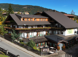 Ein traditionelles, mehrstöckiges Gebäude im alpinen Stil mit einem Satteldach und weitläufigen Balkonen, die mit Blumen geschmückt sind. Im Vordergrund sind Terrassenmöbel zu sehen. Die Umgebung ist von Bergen und Bäumen geprägt, unter blauem Himmel.