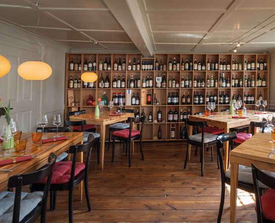 Das Bild zeigt ein gemütliches Restaurant mit Holztischen und -stühlen. Im Hintergrund steht ein Regal, das mit verschiedenen Weinflaschen gefüllt ist. Die Beleuchtung ist warm und bietet eine einladende Atmosphäre. Es sind Tischgedecke mit Gläsern und Servietten zu sehen.