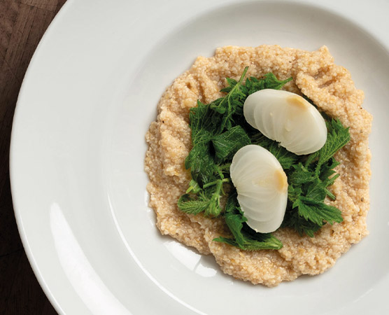 Ein Teller mit cremigem Getreidebrei, der von frischem, grünem Blattgemüse umgeben ist. Obenauf liegen zwei halbierte, gekochte Eier. Das Gericht hat eine ansprechende, rustikale Präsentation auf einem weißen Teller.