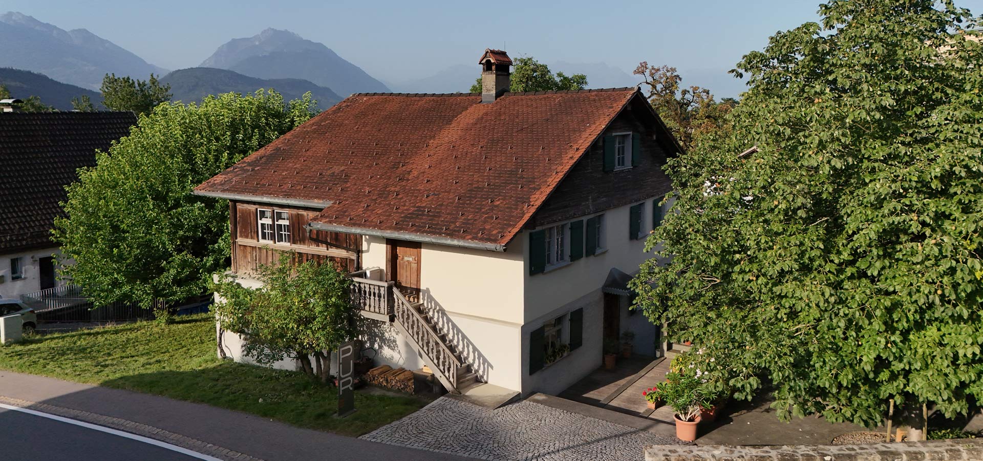 Ein zweigeschossiges, traditionelles Haus mit einem roten Dach und Holzverkleidung an einer Seite. Eine Treppe führt zur Eingangstür, umgeben von grünen Bäumen und Sträuchern. Im Hintergrund sind sanfte Berge zu sehen, während die Sonne den Himmel erhellt.