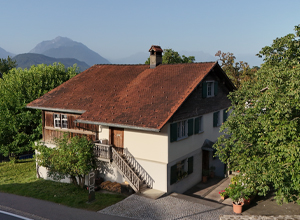 Ein zweigeschossiges Haus im ländlichen Stil mit rotem Ziegeldach und viel Grün umgeben. Die Fassade ist hell gestrichen, mit Fensterläden in einem sanften Blau. Vor dem Haus führen Treppen zur Veranda. Im Hintergrund sind sanfte Hügel und Bäume sichtbar.