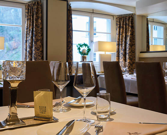 Der Tisch in einem eleganten Restaurant ist festlich gedeckt mit Gläsern, Besteck und einem Menü. Im Hintergrund sind stilvolle Sessel und abgestimmte Vorhänge sichtbar, während das Tageslicht durch große Fenster strömt. Ein gemütliches, einladendes Ambiente herrscht vor.