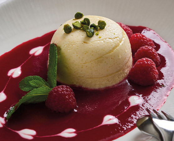Ein souffléartiges Dessert mit einer glatten, hellen Oberfläche liegt auf einem Fruchtspiegel aus roter Beerensoße. Um das Dessert sind frische Himbeeren und ein paar Pistazienstückchen sowie ein grünes Minzblatt platziert. Der Teller hat eine dekorative Anordnung der Soße.