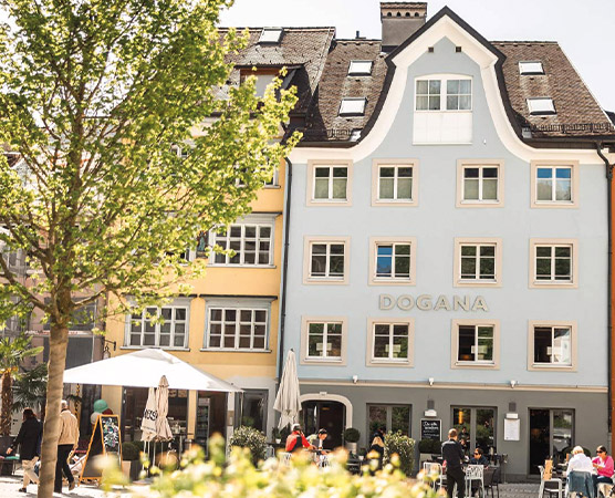 Ein modernes, mehrstöckiges Gebäude mit der Aufschrift „DOGANA“ an der Fassade. Vor dem Gebäude sitzen Menschen an Tischen, während Bäume die Umgebung schattenspendend ergänzen. Es ist ein heller Tag, und die Szene zeigt eine lebhafte Atmosphäre.