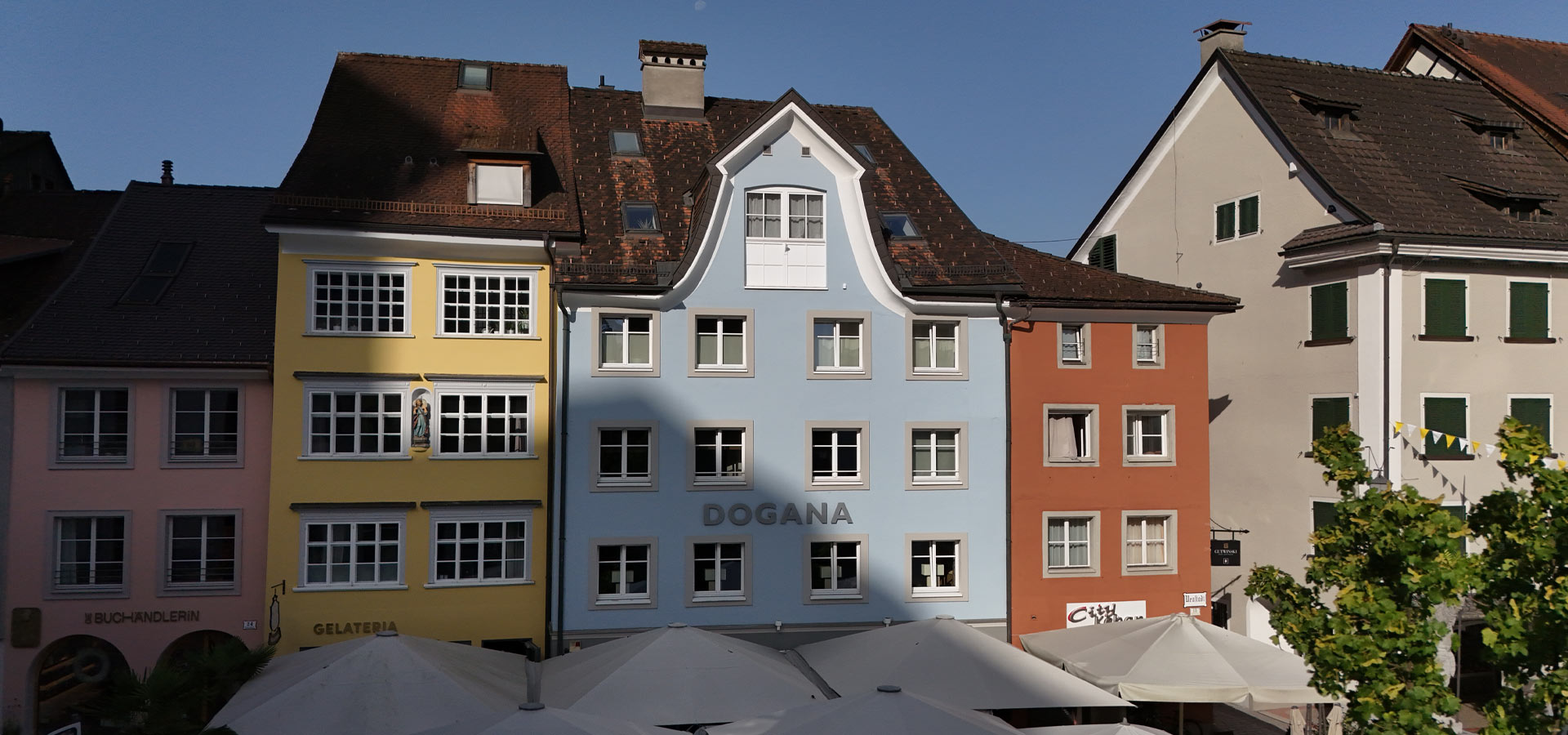 Farbenfrohe Gebäude in einer Stadtansicht, darunter ein blaues Haus mit dem Schriftzug "DOGANA". Die Architektur zeigt typische europäische Fassaden mit Fenstern und schrägen Dächern. Einige Terrassenschirme sind im Vordergrund sichtbar. Der Himmel ist klar und blau.