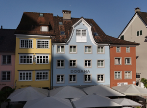 Das Bild zeigt eine Reihe von mehrstöckigen Gebäuden in verschiedenen Farben: ein gelbes, ein blaues mit der Aufschrift "DOGANA" und ein orangefarbenes Gebäude. Im Vordergrund sind Tische mit Schirmen zu sehen, die auf eine Außenterrasse hinweisen. Der Himmel ist klar und blau.