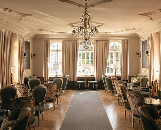 Elegante historische Salon-Lounge mit Kronleuchter, mehreren gepolsterten Sesseln und kleinen Tischen, großen Fenstern mit Vorhängen und hellem Parkettboden.