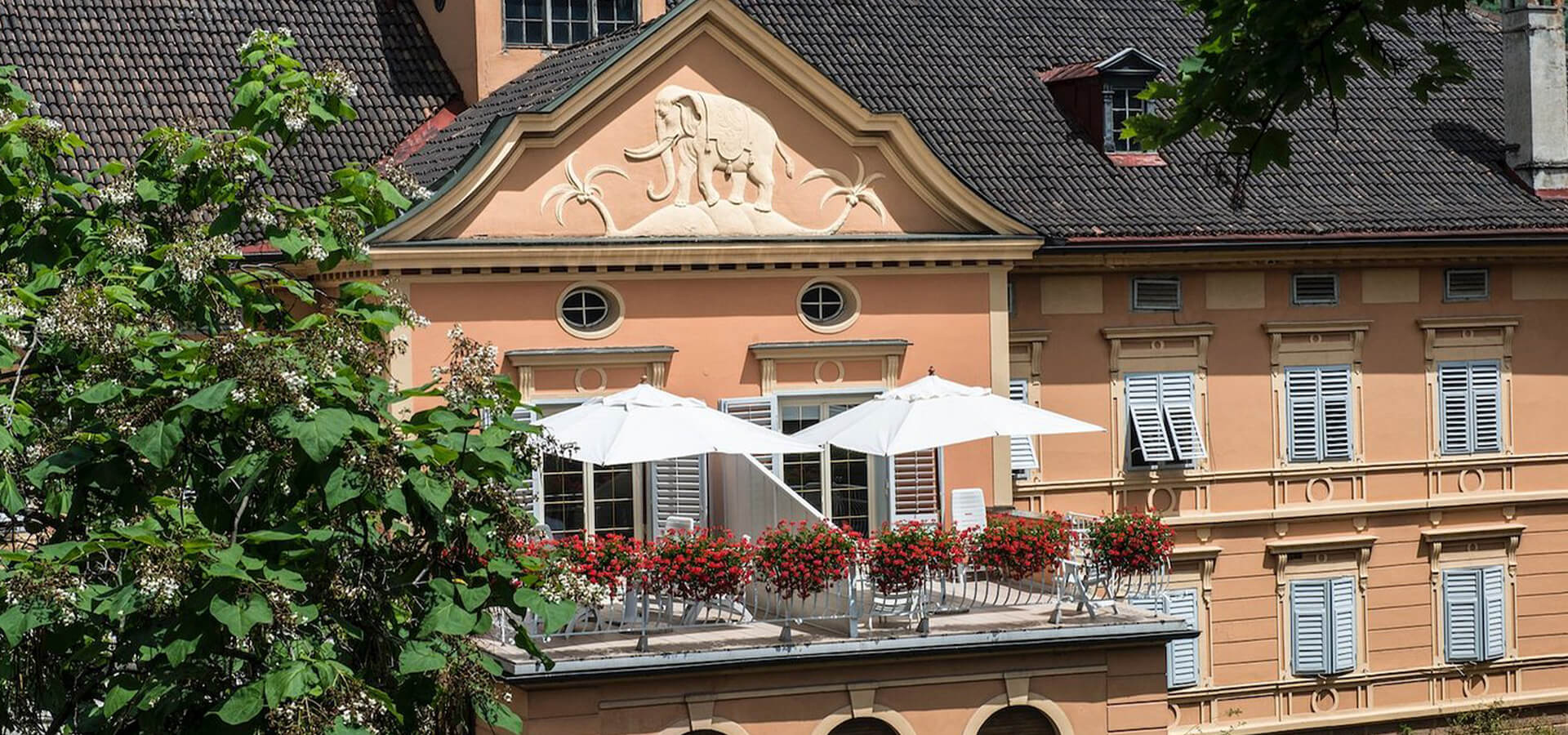 Balkonterrasse eines historischen, hellorangefarbenen Gebäudes mit weißen Sonnenschirmen, roten Balkonblumen, dekorativem Fassadenrelief und geschlossenen Fensterläden.