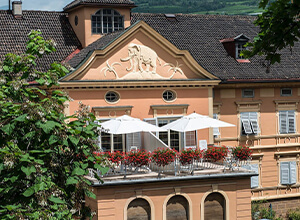 Historisches rosafarbenes Gebäude mit dekorativem Giebel und oberer Terrasse, auf der weiße Sonnenschirme sowie rote Balkonblumen stehen; im Hintergrund bewaldete Hügel.