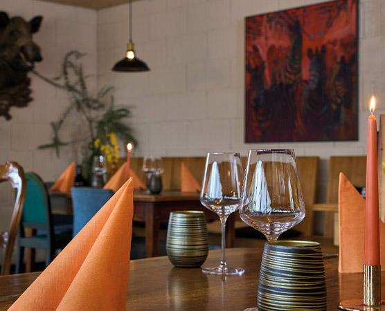 Ein stilvoll gedeckter Tisch in einem Restaurant. Orange Servietten sind zu eleganten Dreiecken gefaltet, während edle Gläser und Kerzen auf dem Tisch stehen. Im Hintergrund ist ein buntes Gemälde und eine Pflanze sichtbar, was eine einladende Atmosphäre schafft.