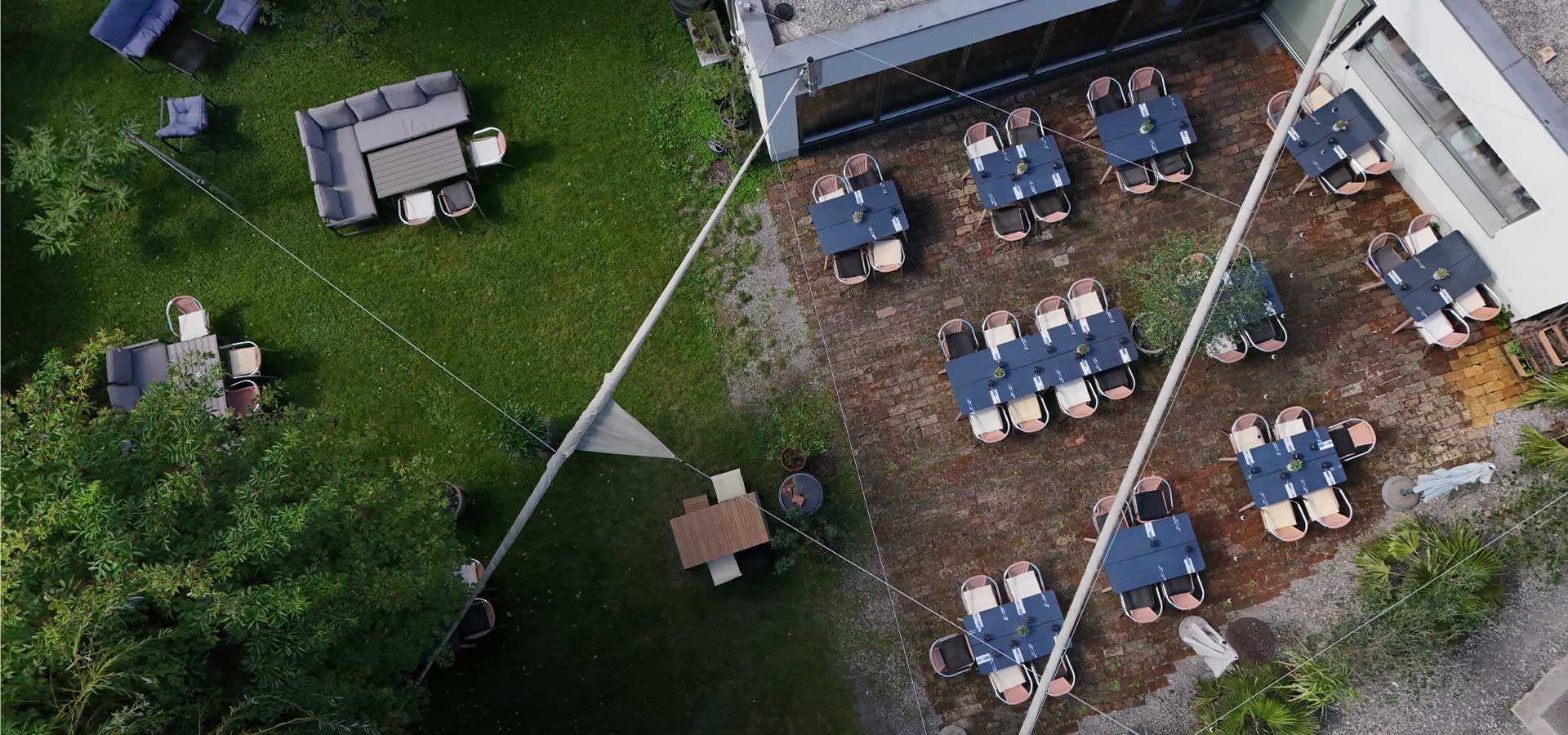 Eine Luftaufnahme eines Restaurantgarten mit mehreren Tischen und Stühlen. Auf der linken Seite sind gemütliche Sitzgelegenheiten, während rechts mehrere Tische auf einem gepflasterten Boden stehen. Ein Sonnensegel und Pflanzen schaffen eine einladende Atmosphäre.