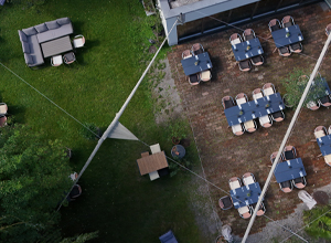 Eine luftige Ansicht eines Gartencafés, das Tische mit Stühlen auf einem gepflasterten Boden zeigt. Im Hintergrund sieht man eine Rasenfläche mit gemütlichen Sitzgelegenheiten. Einige Sonnensegel spannen sich über die Tische, um Schatten zu spenden.