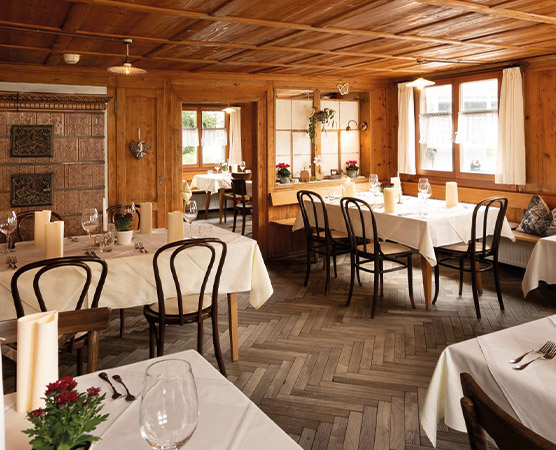 Ein gemütliches Restaurant mit Holzwänden und einem rustikalen Ambiente. Die Tische sind elegant eingedeckt, jeder mit weißer Tischdecke, Gläsern und Kerzen. Pflanzen und dekorative Elemente sorgen für eine einladende Atmosphäre. Fenster lassen natürliches Licht herein.