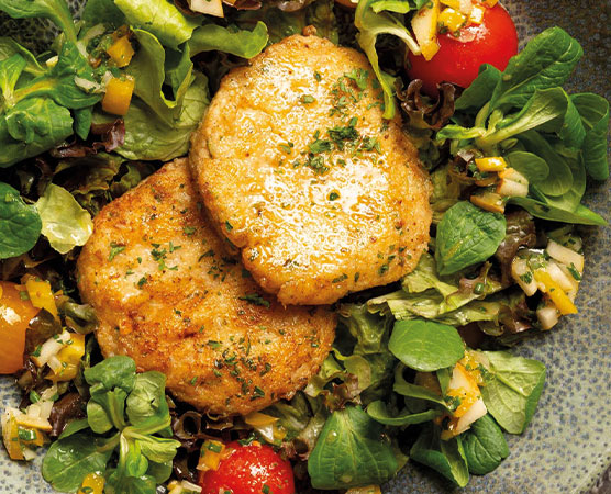 Zwei goldbraune, panierte Fleischlaibchen liegen auf einem bunten Bett aus frischem Salat. Der Salat besteht aus verschiedenen grünen Blattsalaten, Kirschtomaten und einer Zitronen-Vinaigrette, die zusätzlich mit kleinen, bunten Gemüsestücken garniert ist.