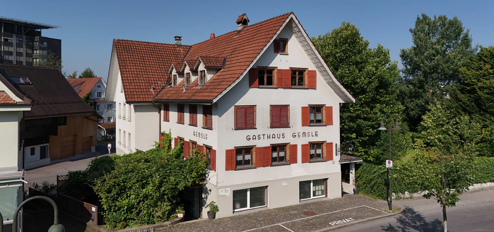 Das Bild zeigt das Gasthaus Gemsle, ein weißes Gebäude mit roten Fensterläden und einem Satteldach. Der Schriftzug "GASTHAUS GEMSL" ist gut sichtbar. Umgeben ist das Haus von Bäumen und Grünflächen, bei klarem blauen Himmel.