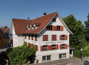 Das Bild zeigt ein mehrstöckiges Gebäude mit einem roten Satteldach und hellen Wänden. An der Fassade steht der Schriftzug „GASTHAUS GIMBLE“. Die Fenster sind mit roten Läden ausgestattet, und das Gebäude ist von grünen Bäumen umgeben. Der Himmel ist klar und blau.