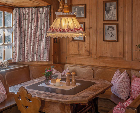 In einem Holzraum befindet sich ein gemütlicher Esstisch mit Kissen und einer Tischdecke. Eine aufwendige Lampe hängt über dem Tisch, der mit einer kleinen Vase und Kerzen dekoriert ist. An der Wand hängen Bilderrahmen mit Fotografien, die dem Raum einen nostalgischen Charme verleihen.