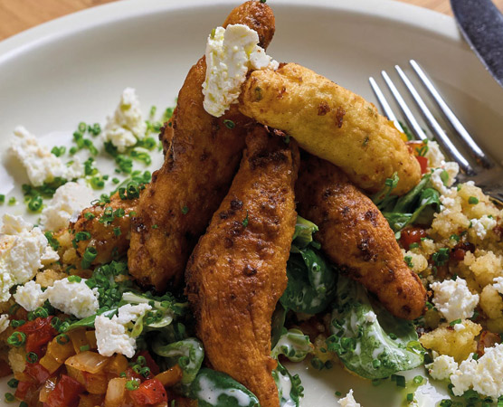 Ein Teller mit frittierten Gemüsestücken, die auf einem Bett aus frischen, grünen Blättern liegen. Um das Gericht herum sind kleine Stücke von zerbröckeltem Käse und buntem Gemüse angeordnet. Eine Gabel liegt neben dem Teller.