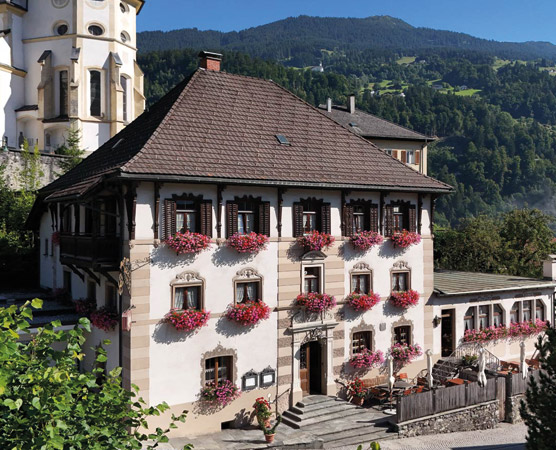 Ein charmantes, historisches Gebäude mit einem Satteldach und dekorativen Blumenkästen an den Fenstern. Es steht in einer malerischen Umgebung mit Bergen im Hintergrund. Vor dem Haus ist ein kleiner Außenbereich mit Sitzplätzen und einer einladenden Atmosphäre.