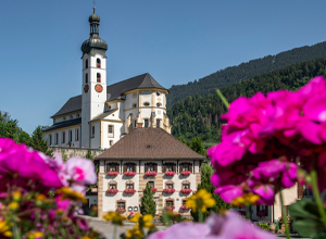 Eine charmante Kirche mit einem hohen Turm steht im Vordergrund einer malerischen Berglandschaft. Im Vordergrund blühen bunte Blumen, die die Schönheit der Umgebung unterstreichen. Die Architektur der Kirche ist traditionell und einzigartig.