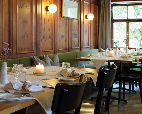 Ein einladender Restaurantbereich mit Holzfurnierwänden. Tische sind elegant gedeckt mit weißen Tischdecken, Gläsern und Besteck. Auf einem Tisch steht eine brennende Kerze, die für eine gemütliche Atmosphäre sorgt. Große Fenster bieten Blick auf die Natur außerhalb.