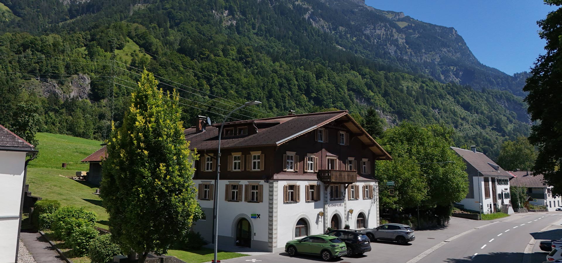 Ein traditionelles Haus mit Holzfassade steht in einer grünen Landschaft, umgeben von Bäumen und Bergen. Auf der Straße parken mehrere Autos. Die Sonne scheint und der Himmel ist klar.