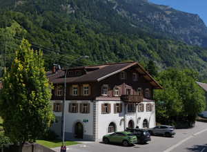 Eine malerische Berghotel-Ansicht zeigt ein traditionelles, dreigeschossiges Haus mit Holzverzierungen, umgeben von üppigem Grün und Bergen im Hintergrund. Parkplätze sind vor dem Gebäude vorhanden. Der Ort strahlt eine einladende, alpine Atmosphäre aus.