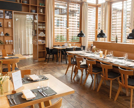 Ein modernes Restaurant mit Holzmöbeln und großen Fenstern. Die Tische sind für Gäste gedeckt, und es stehen Stühle bereit. Im Hintergrund sind Regale mit Produkten. Die Fenster lassen viel Tageslicht herein, was eine helle und einladende Atmosphäre schafft.