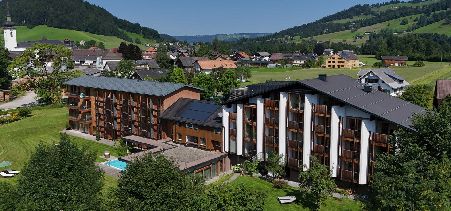 Ein modernes, mehrstöckiges Gebäude mit großen Fenstern und Balkonen steht in einer grünen Landschaft. Im Vordergrund befindet sich ein Schwimmbereich, umgeben von gepflegtem Gras und Bäumen. Im Hintergrund sind einige traditionelle Häuser und sanfte Hügel sichtbar.