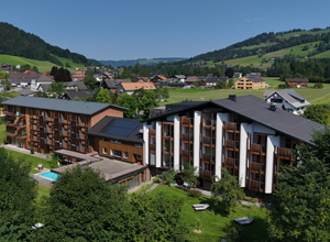 Eine luftige Aussicht auf ein modernes Hotelgebäude mit mehreren Etagen, umgeben von grünen Wiesen und Hügeln. Im Vordergrund sind Bäume und eine Poolanlage sichtbar. Im Hintergrund erstreckt sich eine malerische Landschaft mit weiteren Gebäuden.