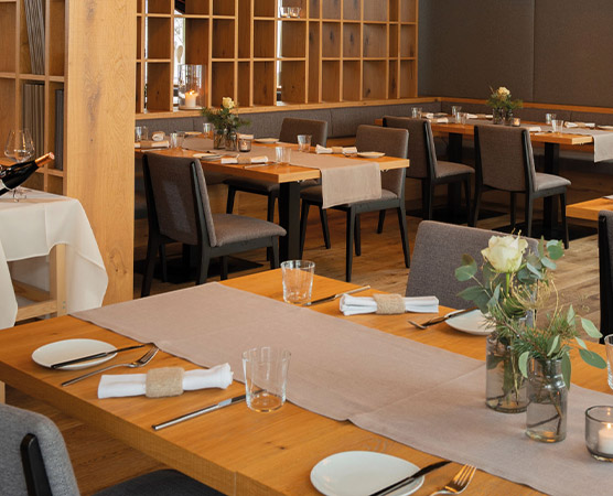 Ein modernes Restaurant mit Holztischen und grauen Stühlen. Die Tische sind gedeckt mit Besteck, Gläsern und kleinen Blumenarrangements. Der Raum wirkt einladend und elegant, ideal für Mahlzeiten mit Freunden oder Familie.